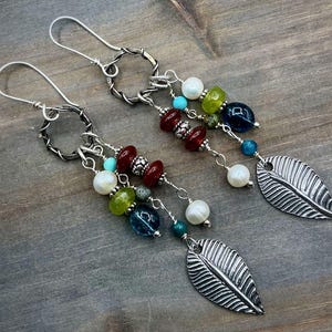 May include: A pair of handcrafted earrings with a nature-inspired theme. The earrings feature silver-toned metalwork, colorful beads in shades of red, green, blue, and white, and a leaf-shaped charm. The earrings are displayed on a wooden surface.