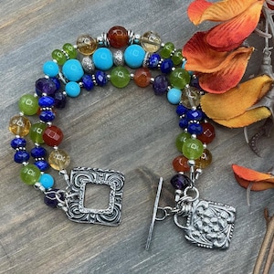 May include: A multi-strand bracelet with a variety of colourful gemstone beads, including turquoise, amethyst, and citrine. The bracelet has a silver-toned clasp with an ornate square design and a matching charm. The beads are strung on a flexible wire.