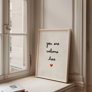 May include: A framed print with the words "you are welcome here" in black script, with a small red heart below. The print is in a light wood frame and sits on a windowsill next to a window.