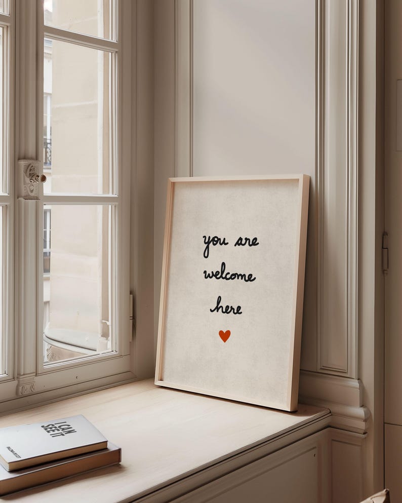May include: A framed print with the words "you are welcome here" in black script, with a small red heart below. The print is in a light wood frame and sits on a windowsill next to a window.