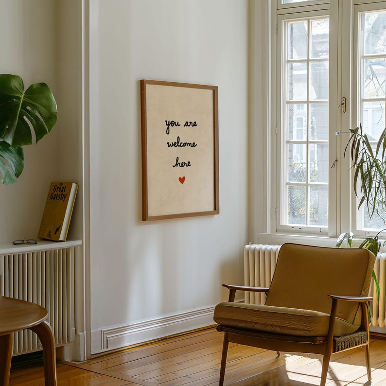 May include: A framed print with the words "you are welcome here" in black script, with a small red heart. The print hangs on a light-coloured wall. A mid-century modern chair and a window are also visible.