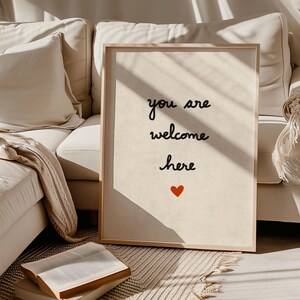 May include: A framed print with the words "you are welcome here" in black script, with a small red heart below. The print has a light beige background and a natural wood frame. The image is set against a neutral-toned sofa.