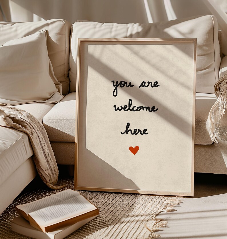May include: A framed print with the words "you are welcome here" in black script, with a small red heart below. The print has a light beige background and a natural wood frame. The image is set against a neutral-toned sofa.