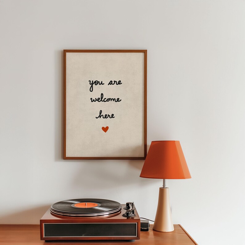 May include: A framed print with the words "you are welcome here" and a red heart. A vintage record player with a vinyl record and an orange lamp are on a wooden surface.