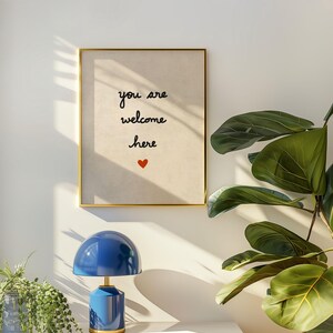May include: A framed print with the words "you are welcome here" and a red heart. A blue table lamp with a gold base sits on a white surface. Green plants are in the foreground.