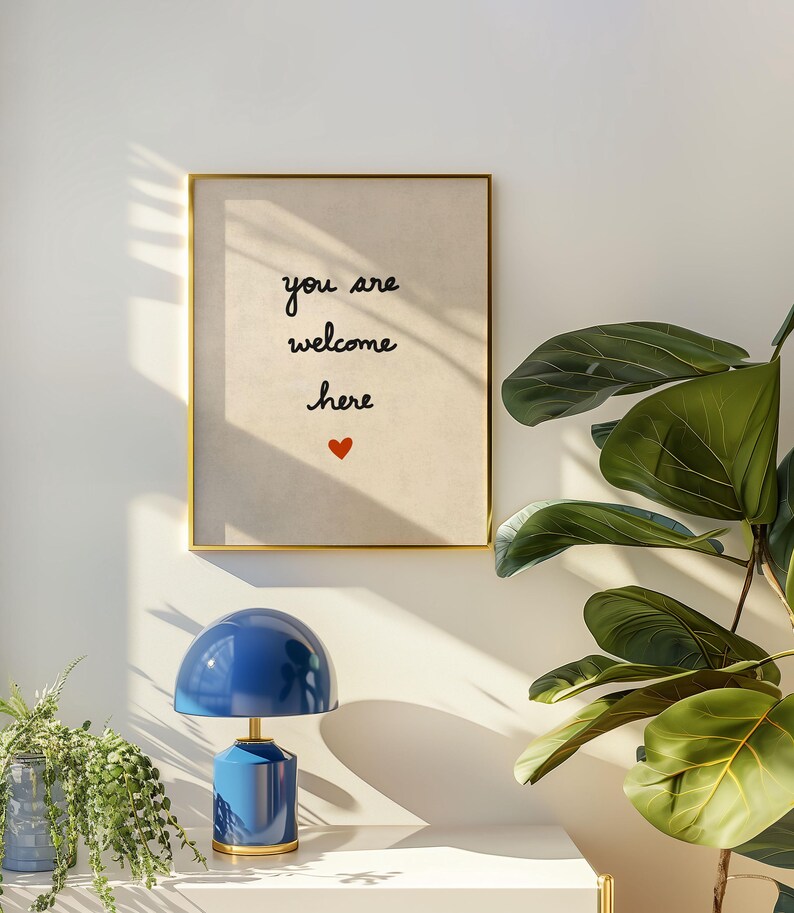 May include: A framed print with the words "you are welcome here" and a red heart. A blue table lamp with a gold base sits on a white surface. Green plants are in the foreground.