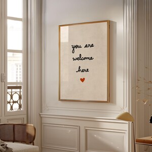 May include: A framed print with the words "you are welcome here" in black script, with a small red heart below. The print has a cream-coloured background and a gold frame, displayed on a white wall in a well-lit room.