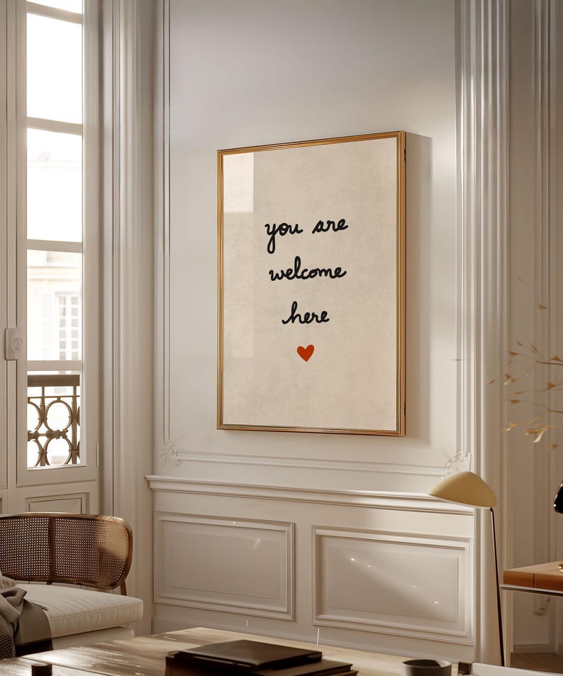 May include: A framed print with the words "you are welcome here" in black script, with a small red heart below. The print has a cream-coloured background and a gold frame, displayed on a white wall in a well-lit room.
