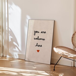 May include: A framed art print with the words "you are welcome here" in black script, a small red heart, and a neutral background. The print is displayed on a wooden floor, near a woven chair.