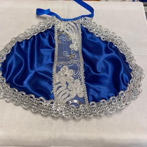 Puede incluir: Una capa de satén azul real con ribete de encaje plateado y detalles bordados. La capa presenta un panel bordado plateado en el centro y una cinta azul para atar. Adecuada para ceremonias religiosas o eventos especiales.
