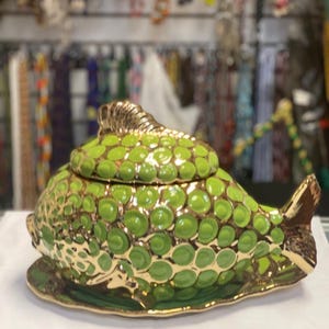 Può includere: Scatola portagioie in ceramica a forma di pesce con coperchio. La scatola è principalmente verde con un motivo strutturato a forma di scaglia e dettagli dorati. Il pesce poggia su un piatto verde abbinato.