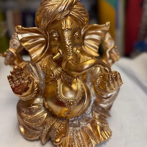 Könnte beinhalten: Eine goldfarbene Statue der hinduistischen Gottheit Ganesha, sitzend mit erhobenen Armen. Die Statue weist komplizierte Details auf, darunter einen Turban, Schmuck und einen Elefantenkopf. Die Figur befindet sich auf einer weißen Oberfläche.