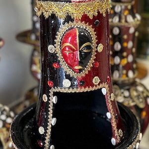 May include: A decorative cylindrical stand featuring a glossy black and red finish, adorned with gold trim, cowrie shells, and a central dual-coloured mask. A small figurine of a child with blue eyes sits atop the structure, wearing a black shirt.