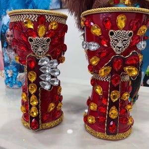 May include: Two ornate red decorative cups with gold trim and leopard face embellishments. The cups are adorned with red, yellow, and clear gemstones. The rims are trimmed with gold rope.