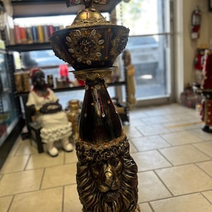May include: A tall, decorative vessel featuring a dark brown and gold finish. The base is sculpted with a prominent lion head, while the upper bowl is adorned with cowrie shells, red beads, and a central face motif, topped with a winged ornament.