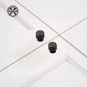 May include: Two matte black cylindrical cabinet knobs with a vertical ribbed texture on the sides. Each knob features a circular top and a narrow base, mounted onto white cabinet doors. The Orlande logo with hammer and ruler icons is visible.