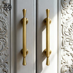 Op de afbeelding: Twee goudkleurige kastgrepen met een verticaal, langwerpig ontwerp. De handgrepen zijn gemonteerd op een lichtgrijze kastdeur. De deur heeft een decoratief, gebeeldhouwd bloemmotief aan de zijkant.