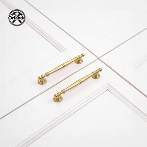 Può includere: Due maniglie dorate per mobili dal design classico, presentate su sfondo bianco. Ogni maniglia presenta una barra centrale con estremità decorative, suggerendo un'estetica vintage o tradizionale. Le maniglie sono probabilmente per cassetti o ante di armadi.