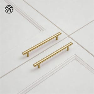 Può includere: Due maniglie dorate per mobili su sfondo bianco. Le maniglie hanno un design cilindrico semplice e moderno. Le maniglie sono orizzontali e parallele. Il logo Orlande si trova nell'angolo in alto a sinistra.