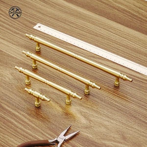 Puede incluir: Cuatro tiradores dorados de diferentes longitudes están dispuestos sobre una superficie de madera. También hay una regla y unos alicates. Los tiradores tienen un diseño clásico con extremos decorativos.