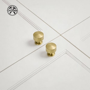 Peut inclure: Deux boutons de meuble dorés avec un design rainuré sont présentés sur un fond blanc. Les boutons sont ronds avec une base cylindrique. Le logo "Orlande" est dans le coin supérieur gauche.
