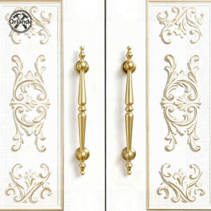 Może przedstawiać: Dwa złote uchwyty do szafek o pionowej, wydłużonej konstrukcji. Uchwyty są zamontowane na białych drzwiach szafki z ozdobnymi, złotymi wykończeniami i dekoracyjnymi akcentami. Na zdjęciu widnieje nazwa marki "Orlande" w lewym górnym rogu.