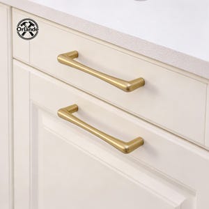 May include: Two gold-coloured cabinet pulls are mounted on a white cabinet. The pulls have a sleek, modern design with a slightly curved shape. The cabinet is part of a kitchen or bathroom set.