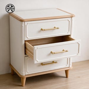 May include: A white bedside cabinet with a light wood trim, featuring three drawers with gold-toned horizontal bar handles. The middle drawer is pulled open, revealing the interior wood construction. The unit stands on four tapered wooden legs.