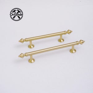 以下が含まれることがあります： 装飾的なフィニアルデザインの2つのゴールドカラーのキャビネットプル。プルは円筒形のバーでまっすぐで、小さな丸いベースに取り付けられています。 Orlandeのロゴは左上にあります。