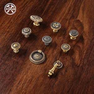 May include: A collection of nine antique brass-toned metal cabinet knobs and pulls featuring intricate floral and geometric embossed patterns. The set includes various circular knob designs and two drop-style pulls with circular backplates.