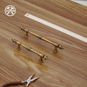 Può includere: Due maniglie per mobili in ottone antico con dettagli decorativi. Le maniglie sono posizionate su una superficie di legno, accanto a un righello e a un paio di pinze. Le maniglie hanno un'estetica vintage.