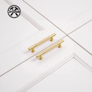 Könnte beinhalten: Zwei goldfarbene Schrankgriffe mit geripptem Mittelteil. Die Griffe sind an einer weißen Schranktür montiert. Das Logo "Orlande" befindet sich in der oberen linken Ecke.