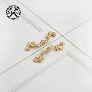 May include: Two ornate, gold-colored cabinet handles with a floral design are displayed against a white background. The handles are curved with decorative detailing, suggesting a vintage or classic style. The handles are likely for furniture or cabinetry.