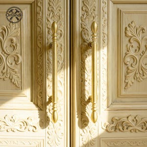 Puede incluir: Primer plano de un par de puertas de madera color crema adornadas con manijas doradas. Las puertas presentan tallas florales detalladas y un logotipo decorativo en la esquina superior izquierda. Las manijas son largas y cilíndricas con extremos redondeados.