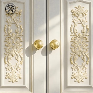 Könnte beinhalten: Nahaufnahme einer weißen Schranktür mit goldfarbener Hardware. Die Tür hat verzierte, erhabene florale Designs und zwei runde, goldfarbene Knöpfe. Der Markenname "Orlande" ist in der oberen linken Ecke sichtbar.