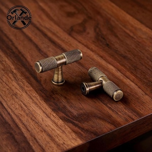Op de afbeelding: Twee T-vormige meubelknoppen met een geborstelde antiek messing afwerking. Elke knop heeft een cilindrische staaf met een getextureerde, gekartelde grip met ruitpatroon aan beide uiteinden en een gladde, taps toelopende centrale voet.