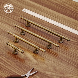 May include: Several antique brass cabinet handles of different sizes. The handles feature a cylindrical bar design with decorative circular bases. A ruler and pliers are also visible on the wooden surface.