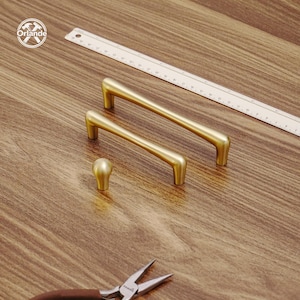 May include: Gold-toned cabinet hardware set, including two bar pulls and a knob. A ruler and pliers are also present on the wooden surface. The pulls have a curved design, and the knob is spherical. The Orlande logo is in the top left corner.