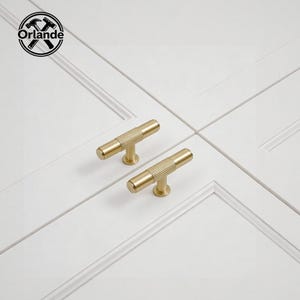 May include: Two gold-colored cabinet pulls with a ribbed, cylindrical design. The pulls are mounted on a white cabinet door. The pulls have a T-bar shape and are made of metal. The logo "Orlande" is in the upper left corner.