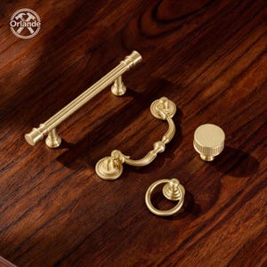 Peut inclure: Un ensemble de quatre pièces de quincaillerie d'armoire en métal doré comprenant une longue poignée cylindrique, une poignée incurvée ornée, un bouton circulaire à bord strié et une petite poignée annulaire, avec le logo Orlande en coin.