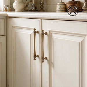 May include: Two vertical antique brass cabinet pulls with a slender, rounded profile and decorative ball-shaped ends, mounted on off-white wooden cabinet doors with a raised panel design. A circular logo with the text Orlande is visible.