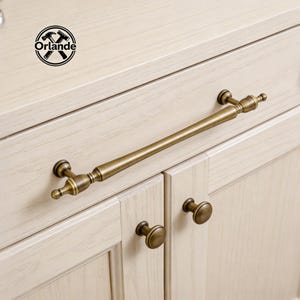Könnte beinhalten: Nahaufnahme von antiken Messing-Schrankbeschlägen. Ein langer, rechteckiger Griff ist über zwei Schranktüren zentriert, mit zwei runden Knöpfen darunter. Die Hardware hat eine Vintage-Ästhetik, mit dem Markenlogo "Orlande" sichtbar.
