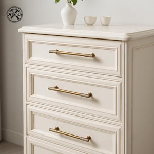 May include: A cream-coloured wooden chest of drawers featuring four rectangular drawers with raised panel detailing. Each drawer is fitted with a long, horizontal, brushed brass metal handle that has a textured, ribbed grip section.