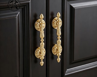 Puede incluir: Dos tiradores de armario verticales de metal dorado con detalles ornamentados y texturizados y placas de montaje circulares fijadas en puertas de armario de madera de color marrón oscuro. El logo Orlande es visible en la esquina superior izquierda.