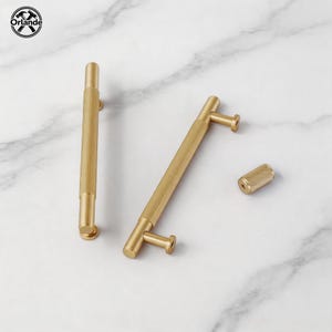 Op de afbeelding: Twee goudkleurige metalen kastgrepen met een textuurrijk, geribbeld middengedeelte en gladde uiteinden, vergezeld van een bijpassende cilindrische knop. Het beslag heeft een geborstelde afwerking en is voorzien van bevestigingspunten.