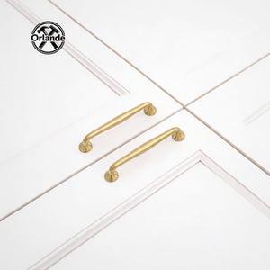 Op de afbeelding: Twee goudkleurige kastgrepen met een gebogen ontwerp zijn gemonteerd op een witte kastdeur. De handgrepen hebben een afgeronde basis en een licht gebogen stang. Het logo "Orlande" staat in de linkerbovenhoek.