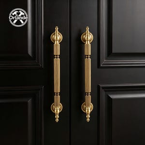 Peut inclure: Deux poignées de porte verticales en laiton avec un design cylindrique cannelé et des embouts arrondis en haut et en bas. Les poignées sont fixées sur une porte sombre à panneaux. Un logo circulaire avec le texte Orlande et des outils croisés est visible.