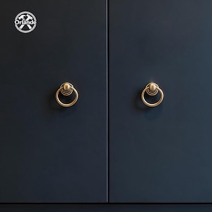 May include: Two matte dark navy cabinet doors featuring circular brass ring pulls. Each door has a centred gold-toned metal handle with a ring design. The top left corner displays a white circular logo with the text Orlande and crossed hammers.