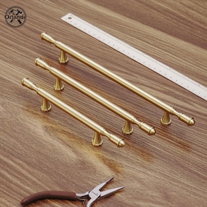 May include: Three gold-coloured cabinet handles of varying lengths, arranged diagonally on a wooden surface. Each handle features a cylindrical bar with decorative end pieces and a central support. A ruler and pliers are also present.