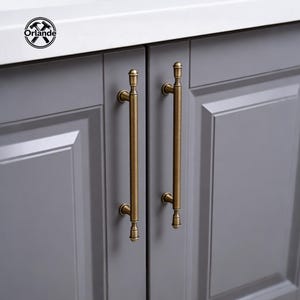 Op de afbeelding: Close-up van twee grijze kastdeuren met lange, messingkleurige handgrepen. De handgrepen zijn verticaal georiënteerd en hebben decoratieve uiteinden. De kastdeuren staan tegen een wit aanrechtblad. Het logo "Orlande" staat linksboven.